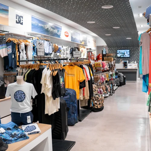 Tienda de ropa de surf con nueva colección en Lanzarote, mostrando camisetas, mochilas y accesorios de moda surf.